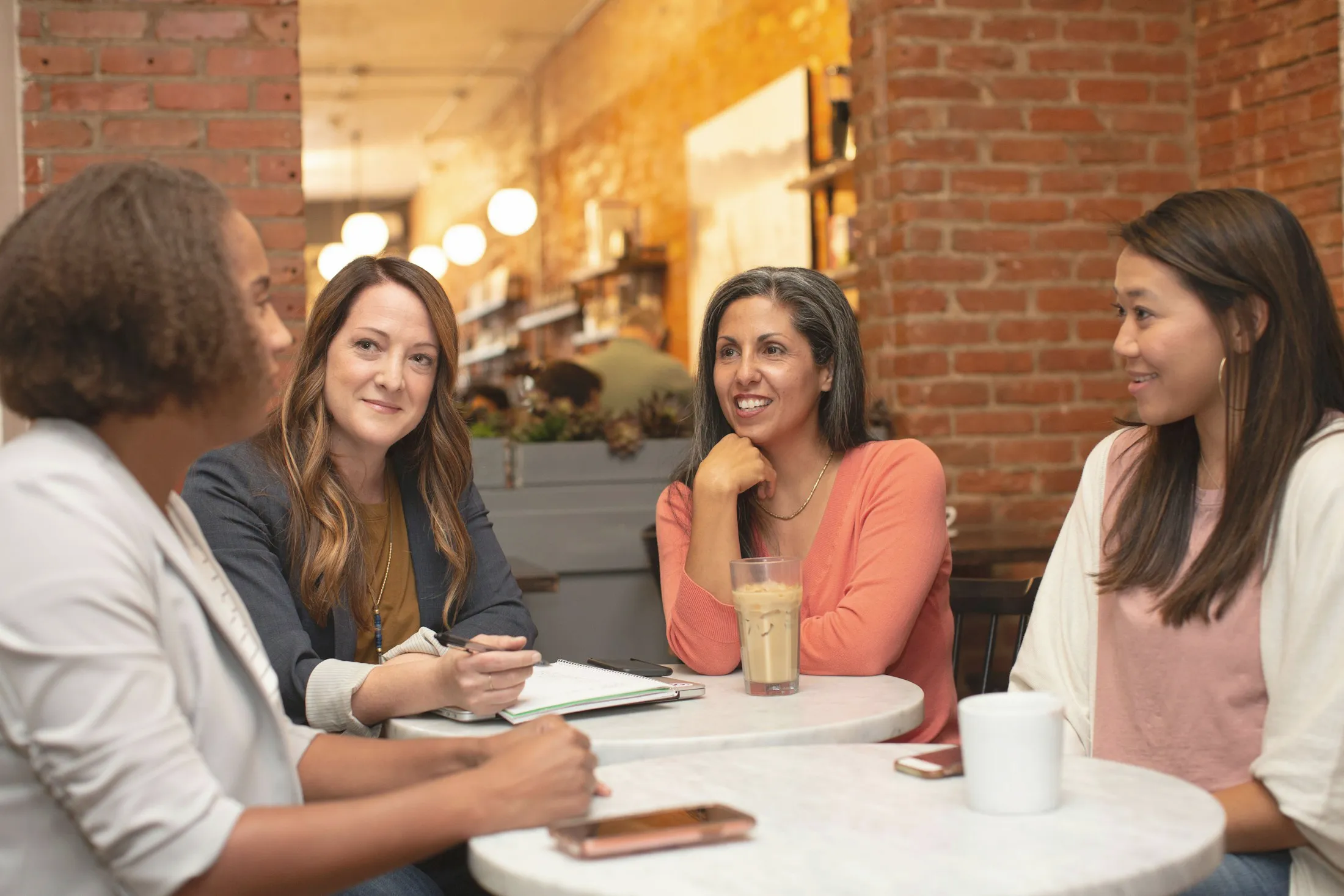 Four women talking around a café table, a notebook open.